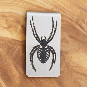 May include: A silver money clip featuring a black spider design. The spider is intricately detailed with a large body and spiky legs. The clip is rectangular with rounded corners and a brushed metal finish. A practical accessory.