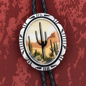 May include: A silver bolo tie with a black leather cord and a round pendant featuring a desert scene with a large cactus and smaller cacti in the background. The pendant has a glass dome over the image.