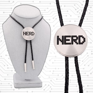 May include: A silver bolo tie with a black braided leather cord. The circular pendant features the word "NERD" in a pixelated font. The tie is displayed on a gray mannequin head against a circuit board background.