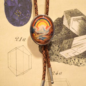 Puede incluir: Un corbatín de cuero marrón con un colgante ovalado de plata que presenta una escena de paisaje colorida con un arcoíris y montañas.