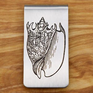 May include: A silver-colored money clip with a detailed black engraving of a conch shell. The shell design is centered on the clip, which has a smooth, brushed metal finish. The background is a light brown wooden surface.