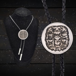 May include: A silver bolo tie with a detailed, circular pendant featuring a textured design. The pendant is attached to a braided black leather cord with silver tips. The tie is displayed on a black mannequin.