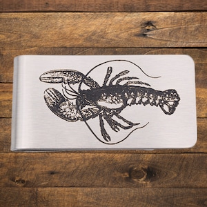 May include: A silver-colored money clip with a detailed engraved illustration of a lobster. The lobster is depicted in black, with its claws and body clearly defined. The clip has a brushed metal finish.