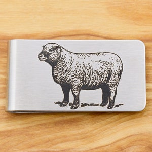 May include: A brushed metal money clip featuring an engraved illustration of a sheep. The sheep is depicted in a detailed, vintage style. The clip is rectangular and has a simple, functional design.