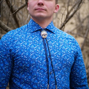 May include: A blue shirt with a white floral pattern and a bolo tie featuring a bison design.