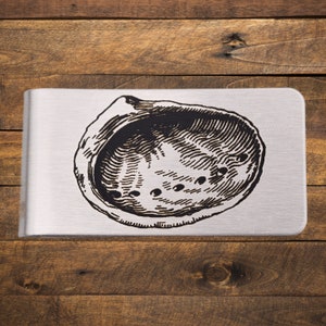 May include: A silver-colored money clip with a black illustration of a shell. The shell design is detailed with lines and small dots. The money clip is rectangular with rounded corners, set against a wooden background.