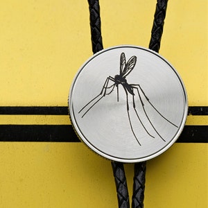 May include: A silver-colored bolo tie with a detailed black mosquito design. The round pendant is attached to a black braided leather cord. The background is yellow with black stripes.