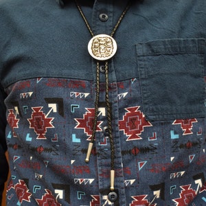 May include: A silver bolo tie with a circular pendant featuring a detailed design. The tie has a black braided cord and silver tips. The bolo tie is worn over a blue button-down shirt with a patterned front panel in shades of blue, red, and white.
