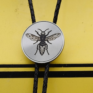 May include: A silver bolo tie with a detailed engraving of a bee on a circular pendant. The pendant is attached to a black braided leather cord. The background is yellow with black stripes.