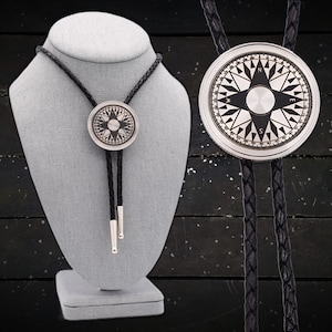 May include: A silver compass bolo tie with a black leather cord. The compass features a black and white design with a silver star in the center. The bolo tie is displayed on a gray mannequin.