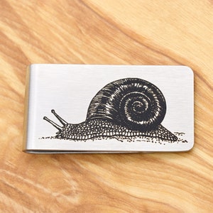 以下が含まれることがあります： 黒い白の彫刻が施された銀色のマネークリップ。カタツムリが右を向いていて、貝殻が細かく彫刻されています。