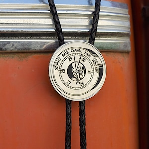 May include: A silver-colored bolo tie with a weather gauge design. The circular pendant features a black and white graphic with the words "Stormy Rain Change Fair Very Dry" around the edge. The tie has a black braided cord.