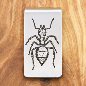 May include: A silver-colored money clip with a detailed black illustration of an ant. The ant design is centered on the clip, which is rectangular with rounded corners. The clip is made of a shiny, brushed metal.