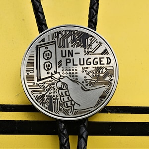 May include: A silver-colored bolo tie with a black braided cord. The circular pendant features an etched design of an electrical outlet, a hand, and the words "UN-PLUGGED". The background is yellow with black stripes.