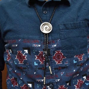 May include: A silver bolo tie with a spiral design pendant. The bolo tie has a black braided cord and silver tips. The person is wearing a blue shirt with a patterned front panel.