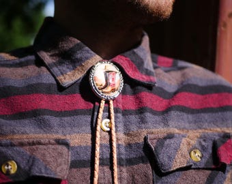 Puede incluir: Una corbata de lazo plateada con un colgante circular con un diseño de bota de vaquero. La corbata tiene un cordón de cuero trenzado y se lleva sobre una camisa a cuadros con rayas burdeos, negras y marrones.