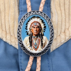 May include: Oval bolo tie with a portrait of a Native American chief. The silver-toned pendant features a detailed image of the chief wearing a feathered headdress and beaded necklaces. The tie has a braided leather cord.