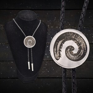 May include: A silver bolo tie with a black braided cord and silver tips. The circular pendant features a detailed fossil design. The tie is displayed on a black mannequin, showcasing the intricate details of the jewelry.