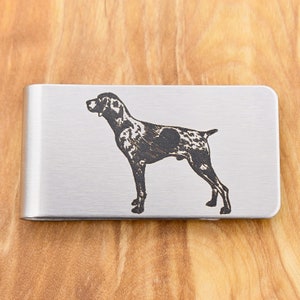May include: A silver money clip featuring a black engraved illustration of a dog, possibly a German Shorthaired Pointer. The dog is shown in profile, with a spotted coat. The clip is made of metal.