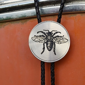 May include: A silver-colored bolo tie with a detailed black bee design on the circular pendant. The tie features a braided black leather cord. The background is a rusty orange color.