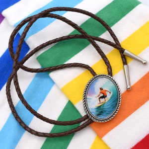 以下が含まれることがあります： 波に乗るサーファーが描かれた銀色の楕円形のペンダントが付いた、茶色の編み込みボロタイ。ペンダントは青、緑、白の背景です。