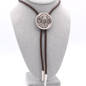 May include: A brown leather bolo tie with a silver round pendant featuring a dog's head engraved on it.