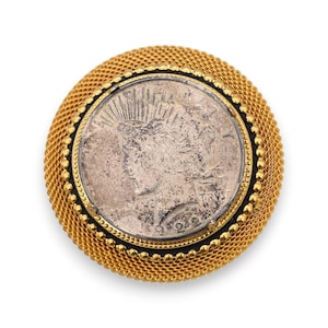 May include: A gold-toned circular brooch featuring a silver coin at its center. The coin has a profile of a woman and is surrounded by a black border, a beaded gold border, and a mesh-like gold outer ring. The brooch is set against a white background.