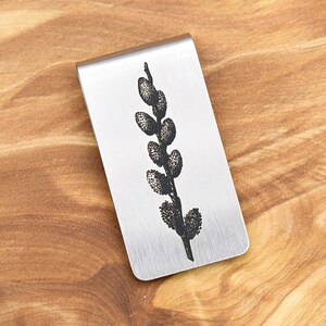 May include: A silver-colored money clip with a detailed black engraving of a pussy willow branch. The clip has a brushed metal finish and a rectangular shape. The design is centered on the clip, showcasing the botanical illustration.
