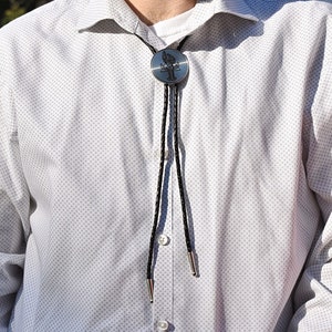 May include: A silver bolo tie with a black cord and silver tips. The bolo tie features a circular pendant with a tree design. The person is wearing a white button-down shirt with a subtle dotted pattern.