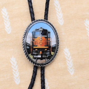 May include: Oval bolo tie with a detailed image of an orange and black train engine on tracks, set against a blue sky. The image is framed with a silver rope-like border. The black braided cord completes the design.
