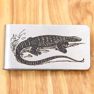 Op de afbeelding: Een zilverkleurige geldclip met een gedetailleerde gravure van een hagedis. De hagedis is in profiel afgebeeld, met zijn lichaam en staart die zich over de clip uitstrekken. Het ontwerp is zwart.