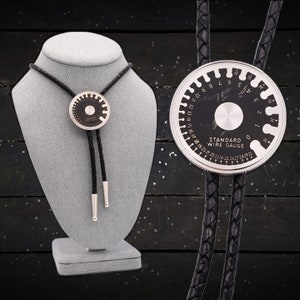 Puede incluir: Una corbata bolo de cuero negro con un colgante de calibre de alambre plateado. El colgante tiene un centro negro con marcas blancas y la inscripción "STANDARD WIRE GAUGE". La corbata se exhibe en un maniquí gris.