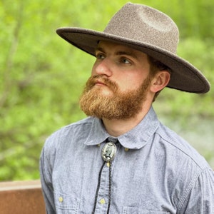Puede incluir: Un hombre con un sombrero fedora gris con una banda negra y una camisa de botones azul claro con una corbata bolo con un diseño de búho.