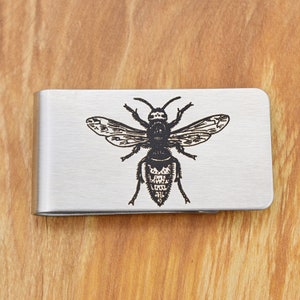 May include: A silver-colored money clip with a detailed black engraving of a bee. The bee design is centered on the clip, showcasing its wings, body segments, and antennae. The clip is rectangular with rounded corners.