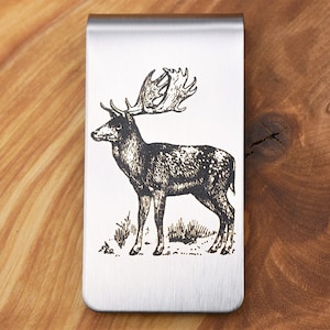 Peut inclure: Un porte-monnaie en argent avec une image gravée au laser d'un cerf avec de grands bois. Le cerf se tient dans un champ herbeux.