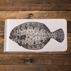 Può includere: Un fermasoldi argentato con una dettagliata illustrazione in bianco e nero di una passera di mare. Il pesce è raffigurato in uno stile artistico dettagliato, con squame e pinne visibili. La clip ha una finitura in metallo spazzolato.