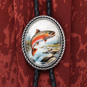 Puede incluir: Un corbatín bolo plateado con un colgante redondo de vidrio que presenta una trucha colorida saltando de un arroyo. El colgante está rodeado por un borde plateado en forma de cuerda.