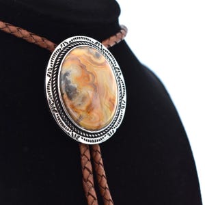May include: A brown leather bolo tie with a silver oval pendant featuring a brown and white agate stone.