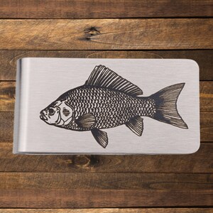 May include: A silver money clip with a detailed black engraving of a fish. The fish is depicted in profile, showcasing its scales, fins, and eye. The clip has a brushed metal finish and a rectangular shape.