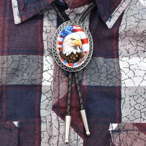 May include: A bolo tie with a silver oval pendant featuring an American bald eagle with an American flag in the background. The bolo tie has black leather cords and silver tips.
