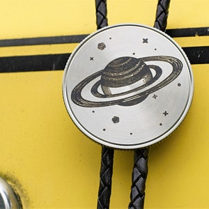 May include: A silver bolo tie with a detailed engraving of Saturn and its rings, surrounded by stars. The pendant is attached to a black braided leather cord. The background is a bright yellow surface with black stripes.