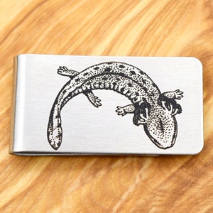 Op de afbeelding: Een zilverkleurige geldclip met een gedetailleerde zwarte illustratie van een salamander. De salamander, met zijn gevlekte patroon en kleine ledematen, is over het oppervlak van de clip gebogen. De clip is rechthoekig met afgeronde hoeken.