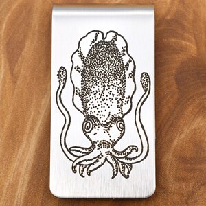 May include: A silver-colored money clip featuring a detailed black ink illustration of a squid. The squid design is centered on the clip, with the tentacles and body clearly visible. The clip is rectangular with rounded edges.