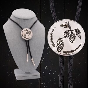 May include: A silver bolo tie with a black braided leather cord. The circular pendant features a detailed engraving of hops and leaves. The cord ends have silver tips. The tie is displayed on a gray mannequin.