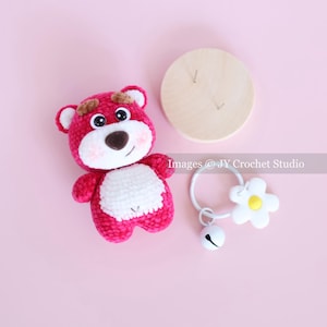 May include: A handmade, pink crochet bear with a white belly and brown accents. A wooden circle and a white baby rattle with a flower and bell are also present. The items are on a pink background.