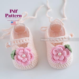 May include: A pair of light pink crochet baby shoes. Each shoe has a pink flower with a pearl centre and a green leaf accent. The shoes feature a strap with a pearl button closure and a delicate tie.
