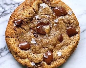 May include: A close-up of a chocolate chip cookie with sea salt on top. The cookie is golden brown and has a soft, chewy texture.