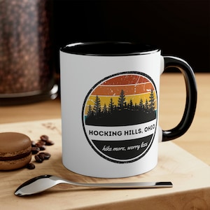 May include: White ceramic coffee mug with a black rim and handle. The mug features a retro-style graphic of a forest with a sunset behind it. The text "Hocking Hills, Ohio" and "hike more, worry less" is printed on the mug.