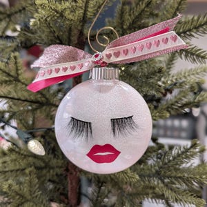 May include: A white glitter Christmas ornament with a pink glitter ribbon and a painted design of eyelashes and red lips.