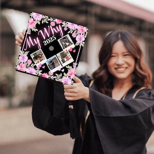 May include: A black graduation cap decorated with pink and purple floral accents and the words "My Why 2025." The cap features four small photo prints. The person holding the cap is wearing a black graduation gown.